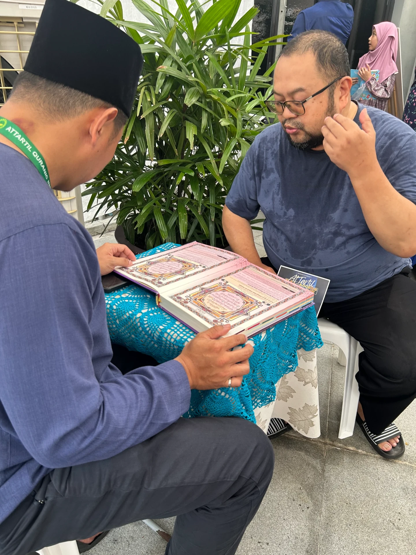 A teacher reading an illuminated Qur'an with a visitor at the booth