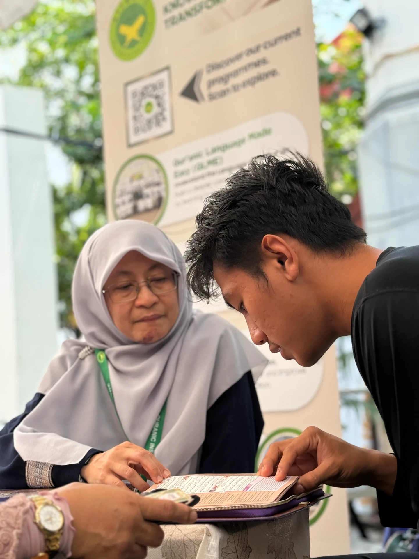 A young man reading the Qur'an with an AtTartil QI teacher beside the booth banner